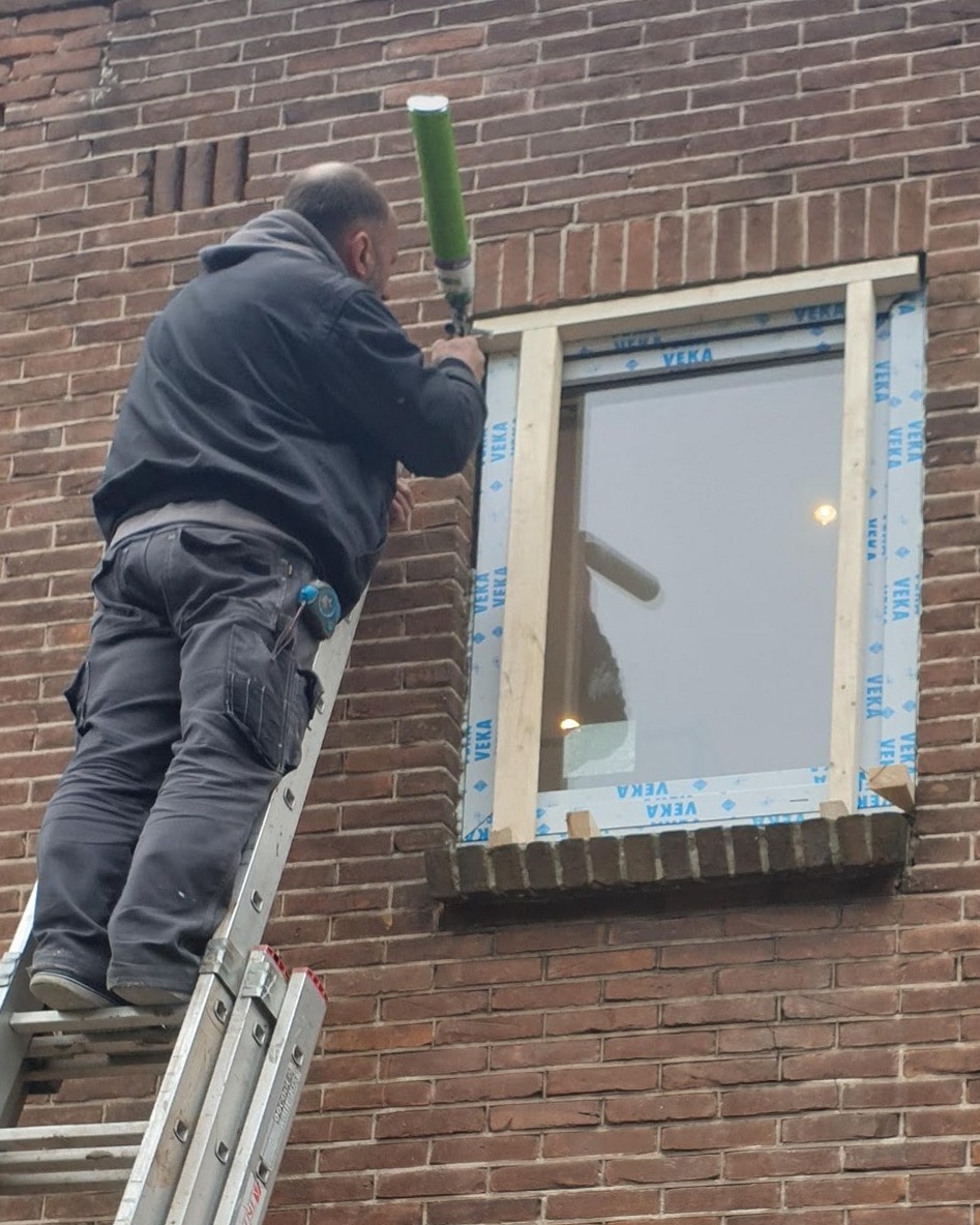 Monteur op een ladder schuimt een nieuw kunststof kozijn in een bakstenen gevel