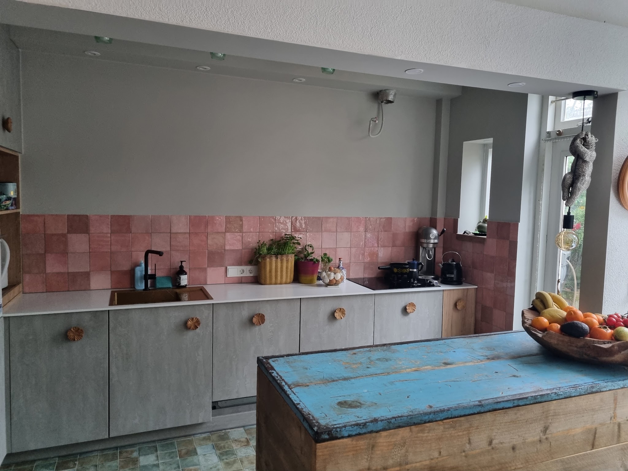 Keuken met roze zellige-achtige wandtegels, betonlook fronten, houten grepen en een kookeiland van blauw sloophout